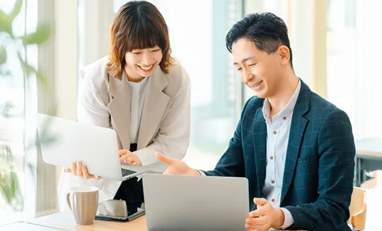 オフィスでノートパソコンを囲み笑顔で打ち合わせをする男女。ITサービスの円滑な導入と活用をイメージ